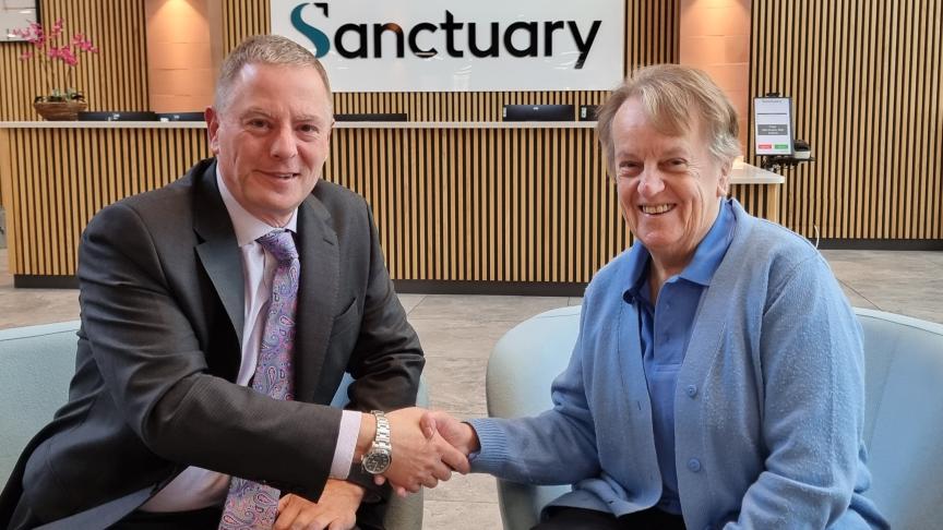 Two people are seated in modern chairs in a reception area, shaking hands. Behind them is a wooden-panelled wall with a large sign that reads “Sanctuary.”