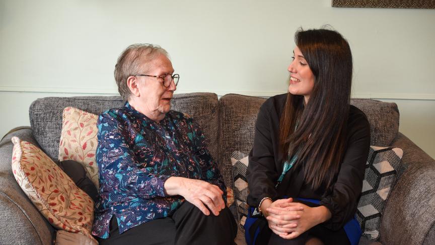 senior woman and a younger woman are sitting together on a cozy gray sofa, smiling and engaging in conversation. The older woman, wearing glasses and a patterned blouse, appears joyful, while the younger woman, dressed in a black top, is laughing. Decorative pillows are arranged on the sofa, and the background features soft lighting and a hint of wall decor, creating a warm and inviting atmosphere.