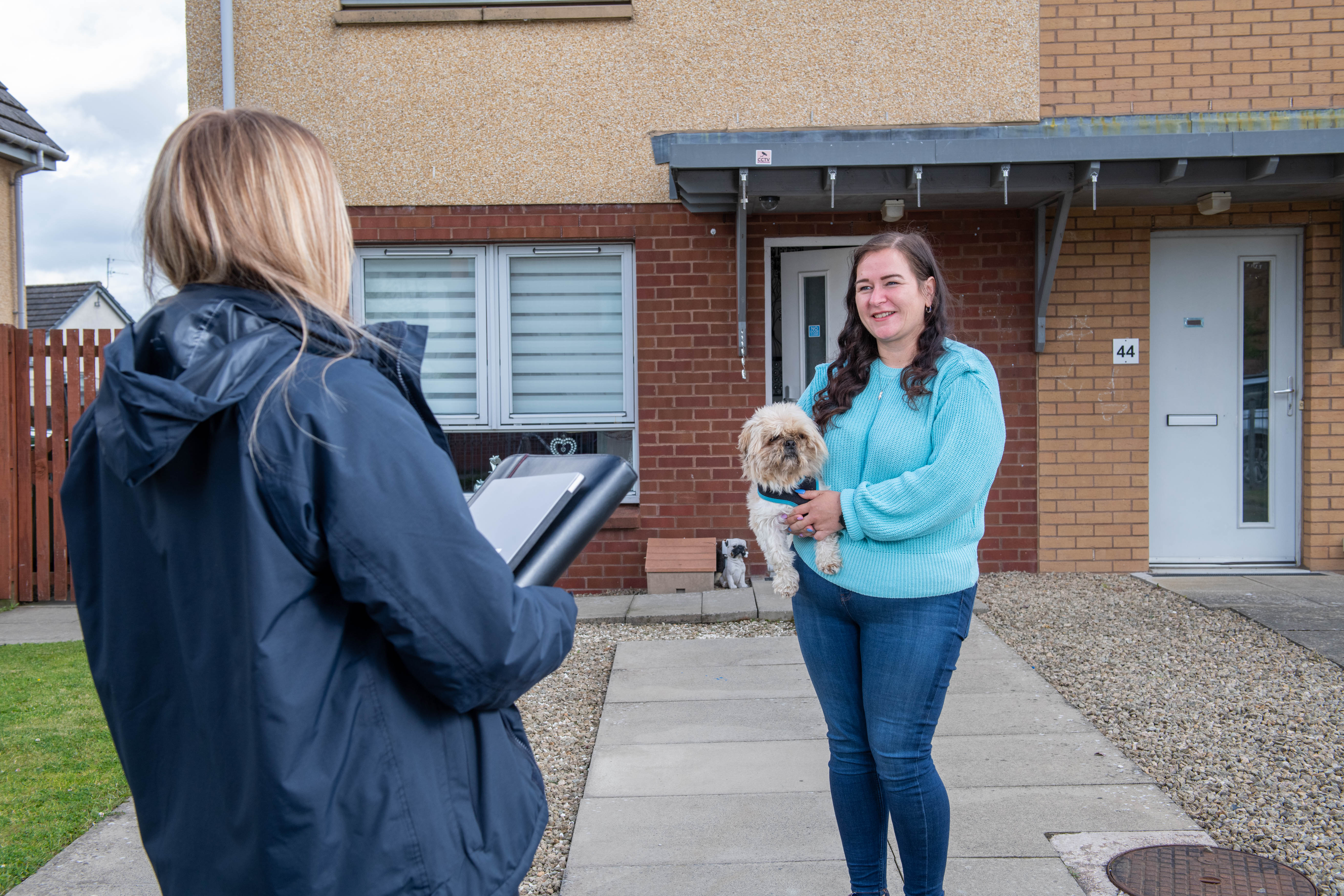 A person outside the front of their home holding a small dog facing another person who looks as though they are working for an organisation with a folder in their hand