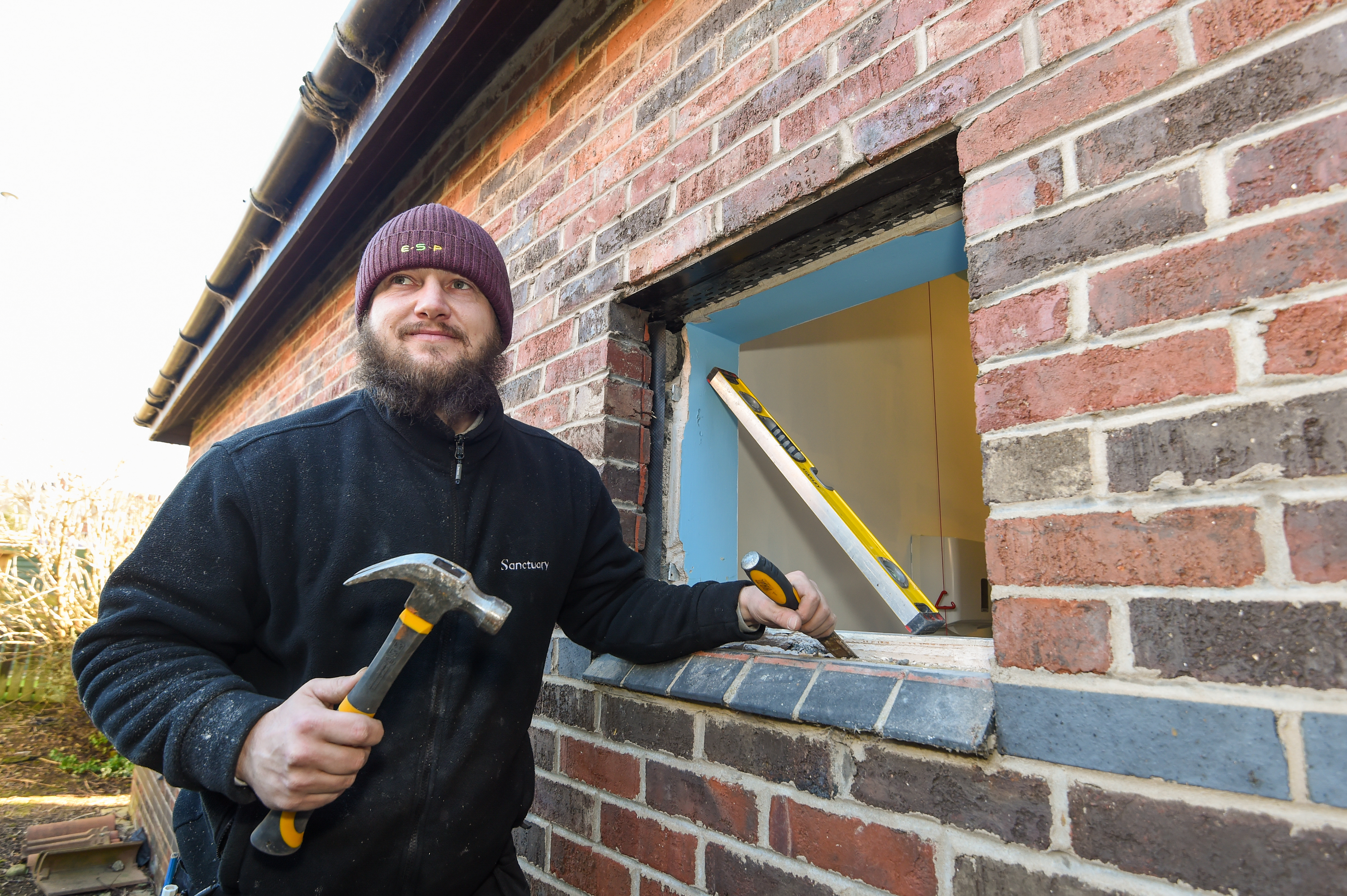 A man in a navy Sanctuary-branded fleece holds a chisel and hammer in front of a window where the double-glazing has been removed