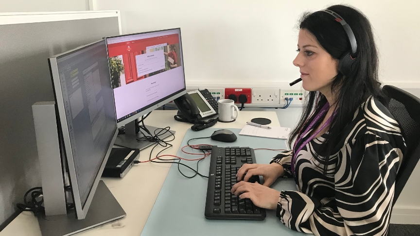 A member of Sanctuary Supported Living staff sat at a desk working on a computer whilst wearing a telephonw headset