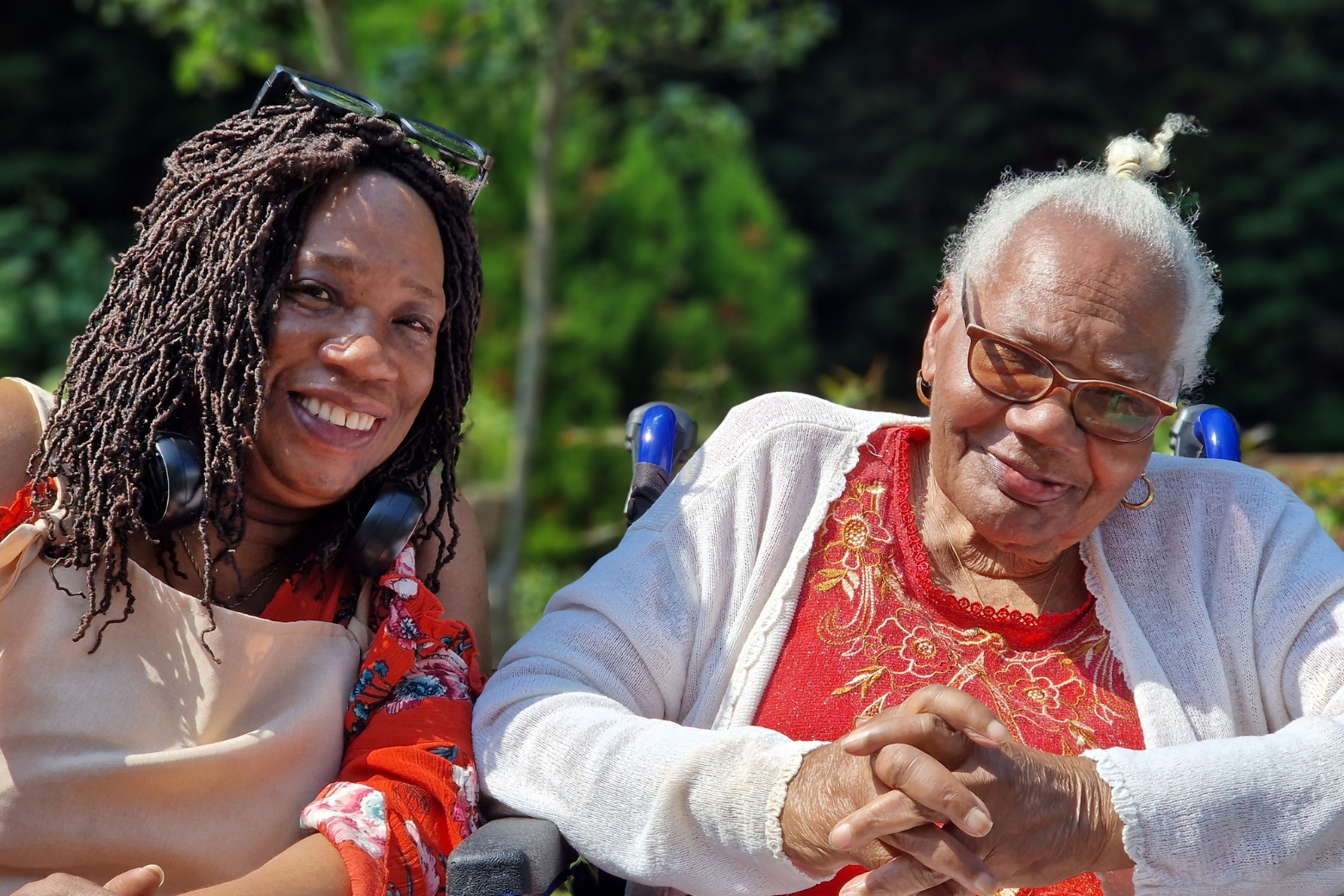 A mother and daughter sat next to each other smiling at the camera, the older woman is sat in a wheelchair