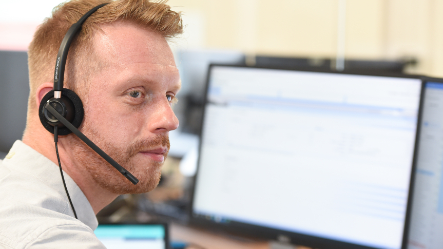 A ginger haired man with a beard wearing a phone headset. There are computer screens on a desk visible in the background.