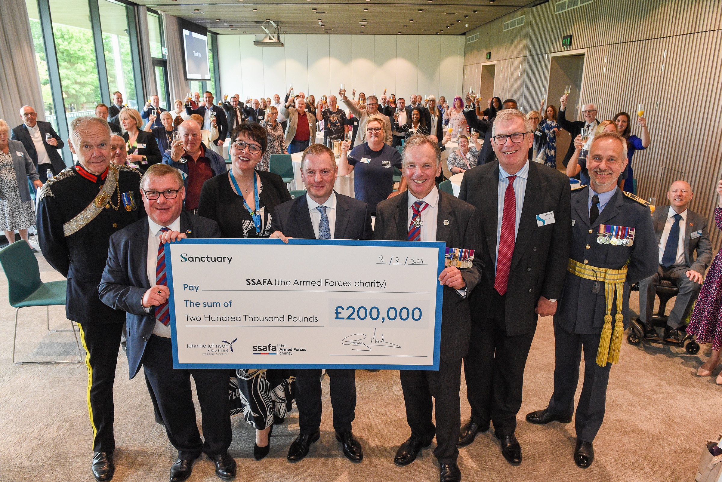 A large group of people standing in a room raising their glasses in celebration. In the forefront of the picture are six males and one female holding a large cheque for £200,000 from Sanctuary to the Armed Forces charity. Some of the men are wearing military medals.