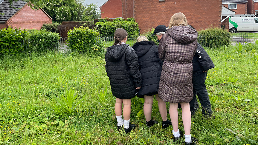 A woman and school children standing in a greasy field with redbrick houses in the background