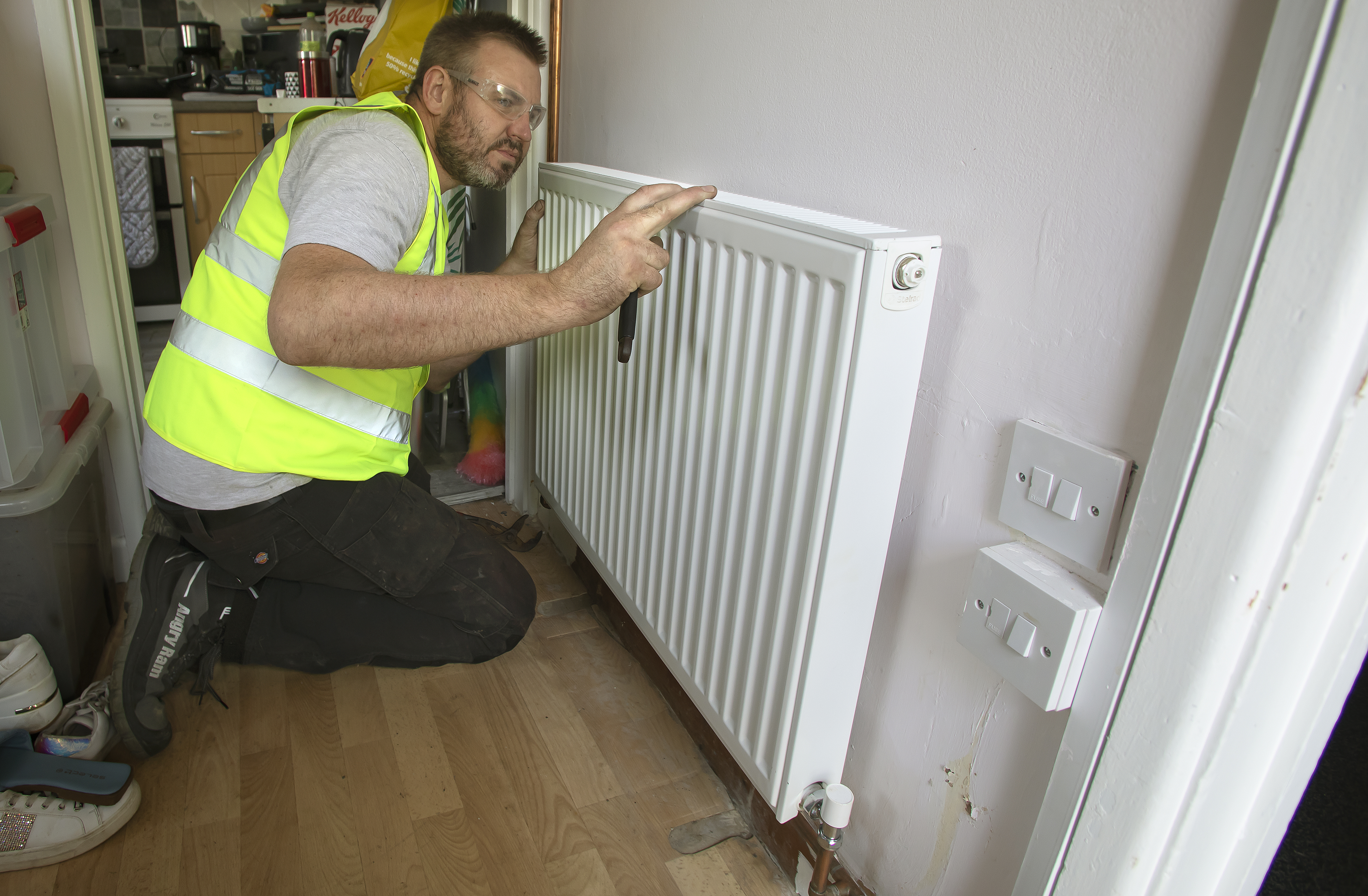 A man wearing a yellow high visibility jacket and protective goggles kneeling in a hallway working on a radiator