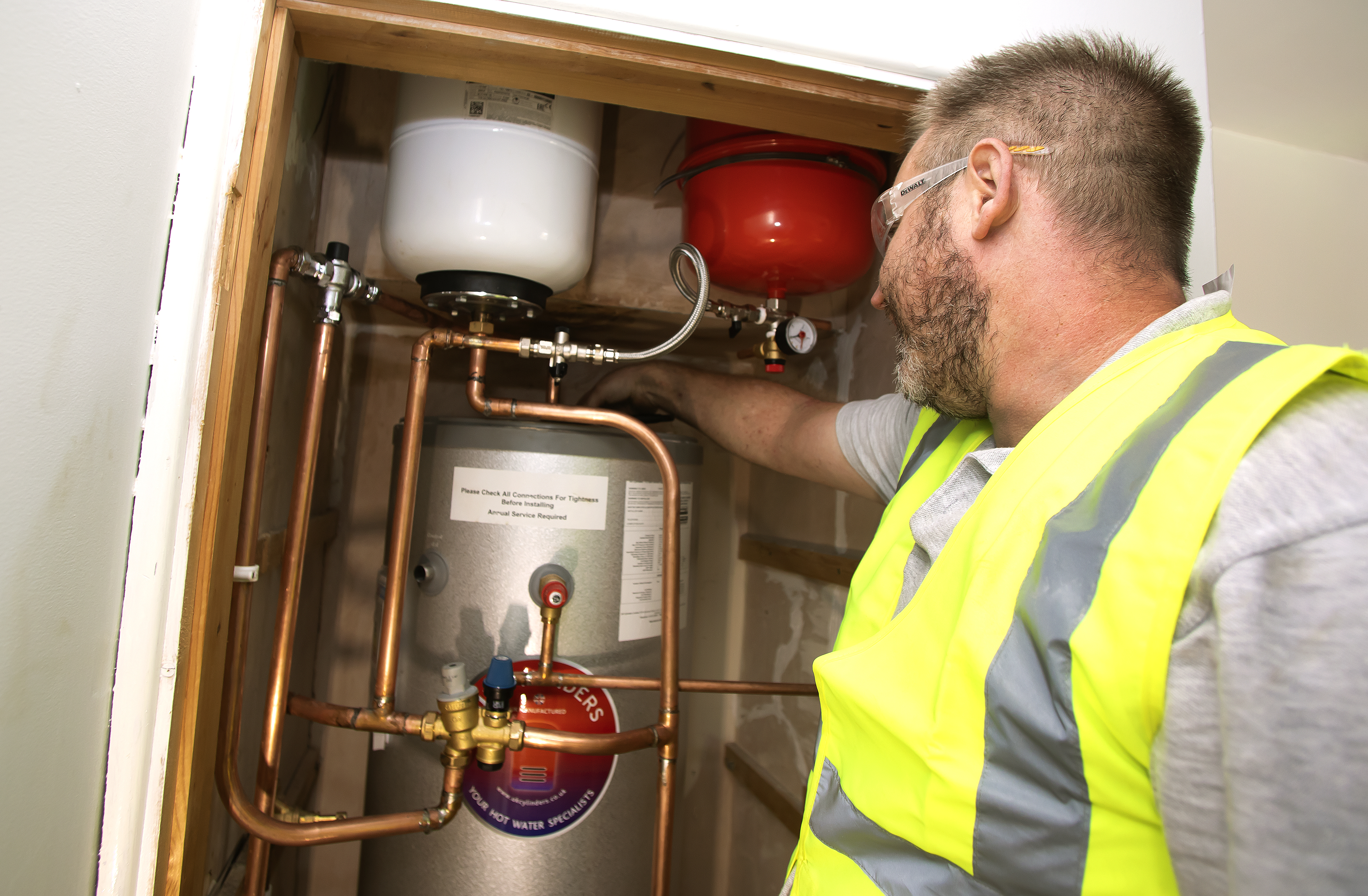 A technician wearing a safety vest and glasses is inspecting a water heater installation. The water heater is cylindrical and metallic, with various copper pipes connected to it. The technician is pointing at a component on the heater, which is mounted in a wooden enclosure. The background shows unfinished walls, indicating that the area is still under construction.