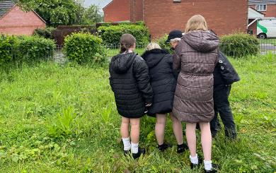 A woman and school children standing in a greasy field with redbrick houses in the background