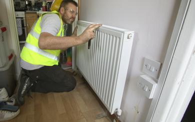 A man wearing a yellow high visibility jacket and protective goggles kneeling in a hallway working on a radiator