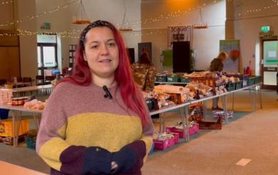 A woman standing in a large room with tables behind her stacked with food and produce