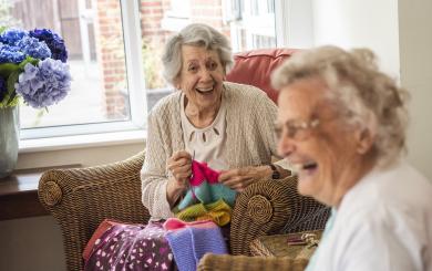 Two elderly women sitting in wicker chairs and laughing whilst knitting with very bright coloured wool