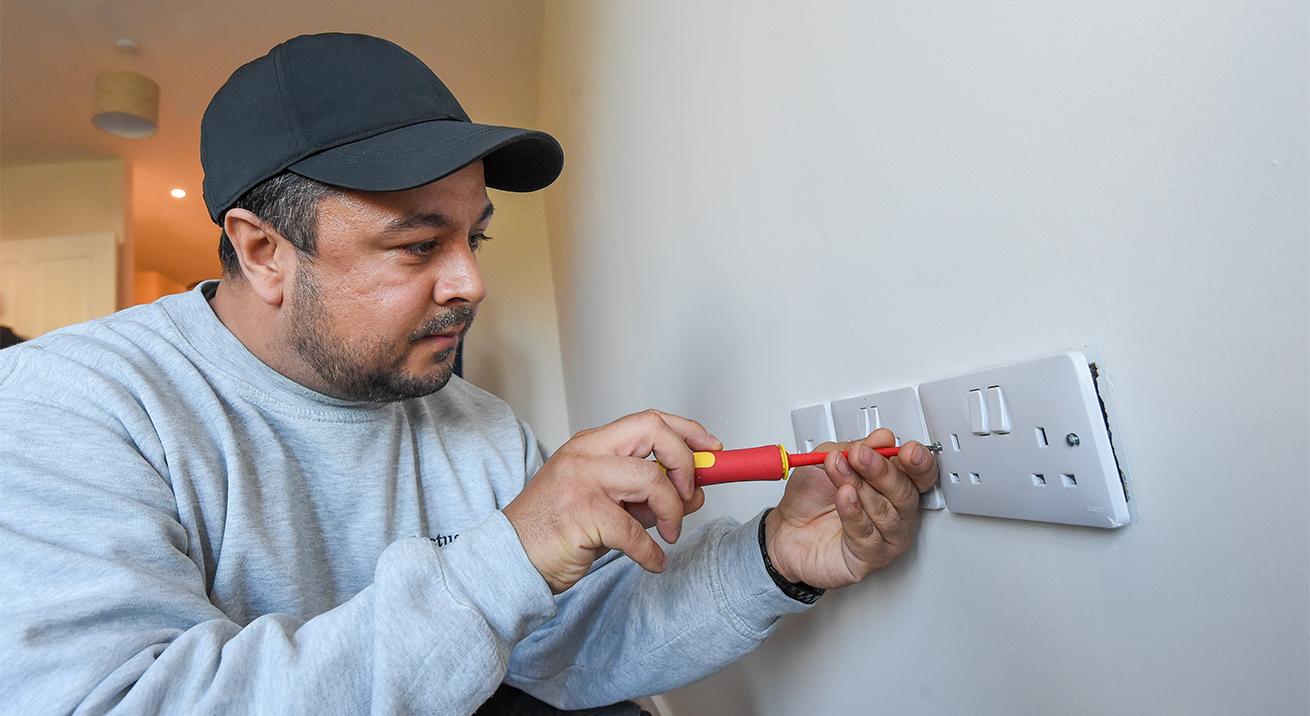 A sanctuary maintenance worker wearing a grey jumper and black baseball cap using a red screwdriver to screw in a white plug socket into a white wall