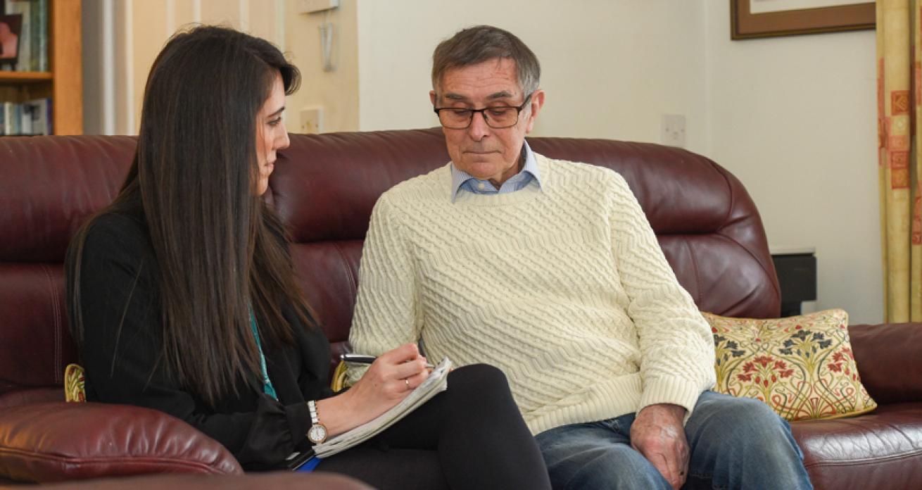 A woman sitting on a leather sofa wearing a Sanctuary lanyard and writing in a notebook looking at an older man sitting next to her on the sofa