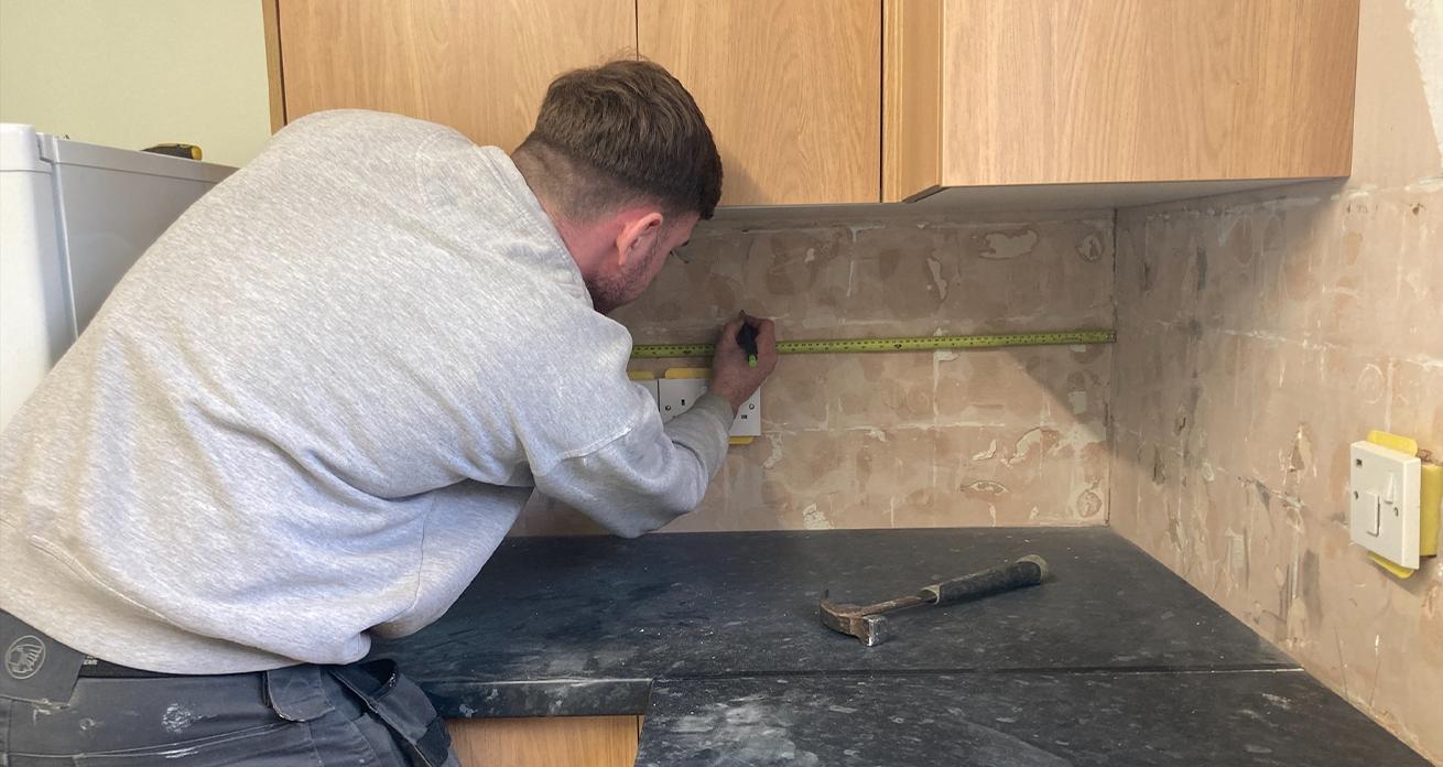 A man wearing grey trousers and jumper uses a measuring tape to measure a distance on a tiled wall with wooden cupboards above and a black counter below