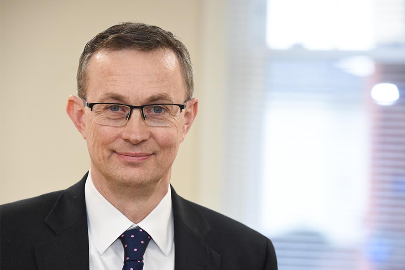 A man with brown hair wearing glasses, a black jacket, white tie and pink spotted tie looking at the camera and smiling 