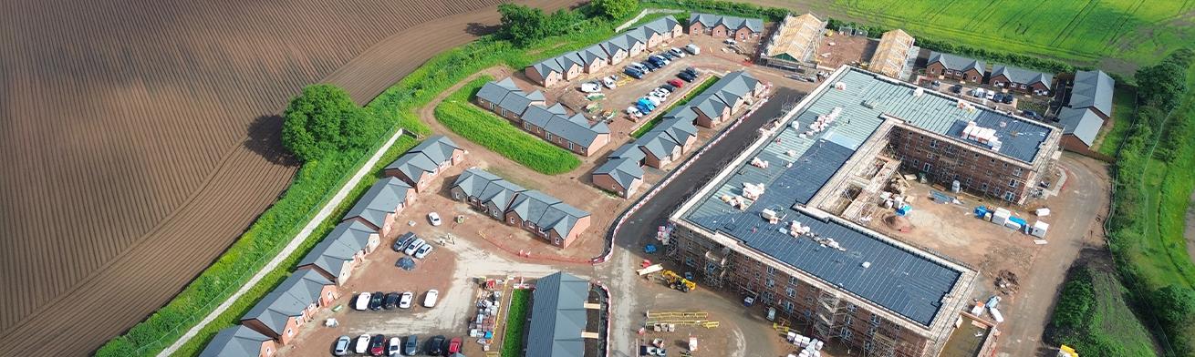 An aerial view of a housing development consisting of semi-detached houses and a block of flats. There is a green field on the right of the development and a ploughed field on the left of the development