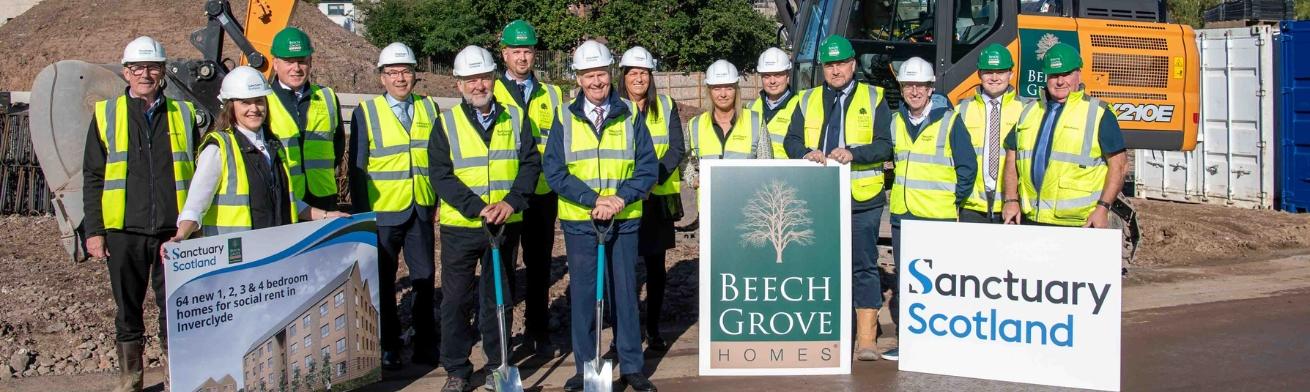 A group of 14 people wearing high vis and hard hats stood in front of a housing development site on a sunny day