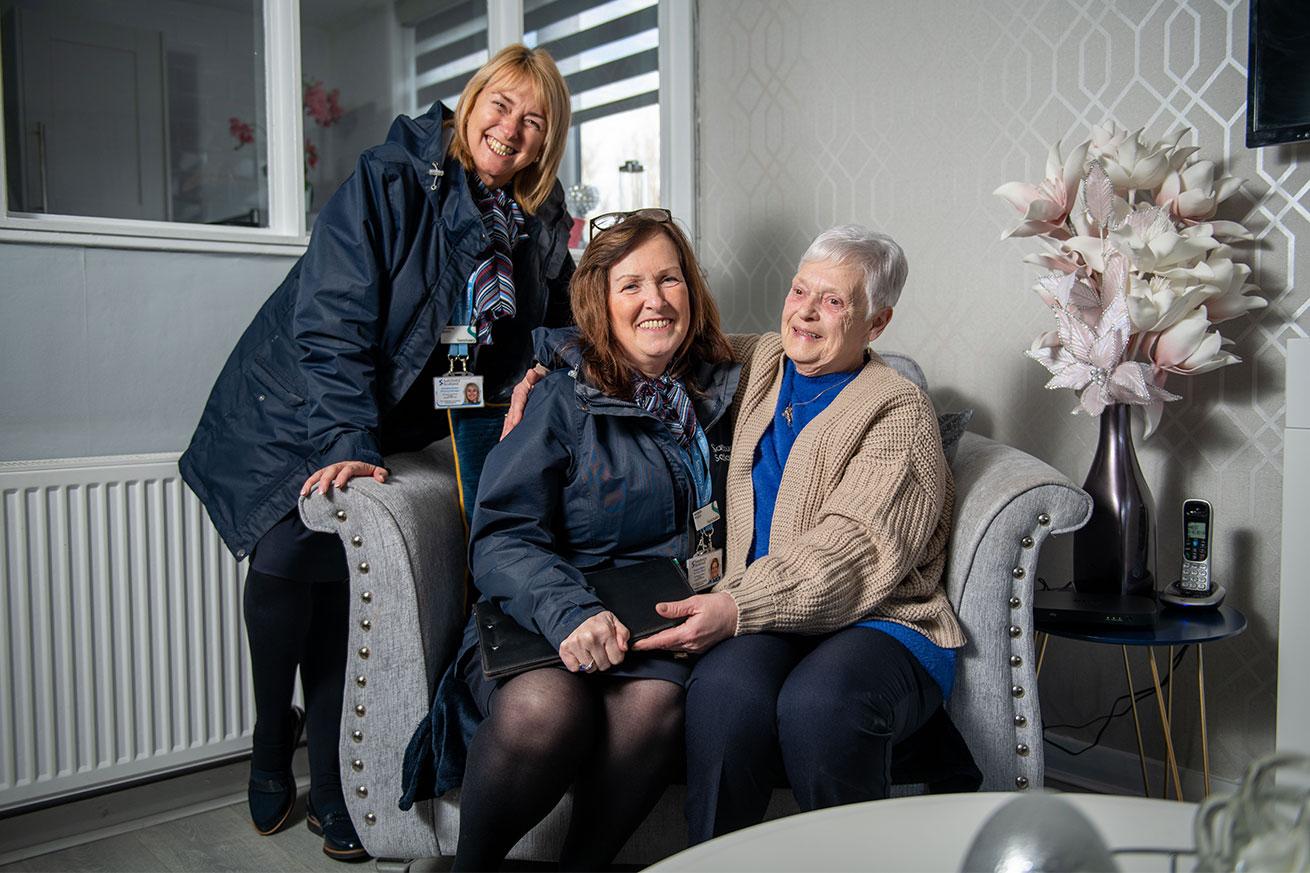 Two Sanctuary Scotland Housing Officers sitting in a living room with an elderly resident who has their arm around one of the housing officers