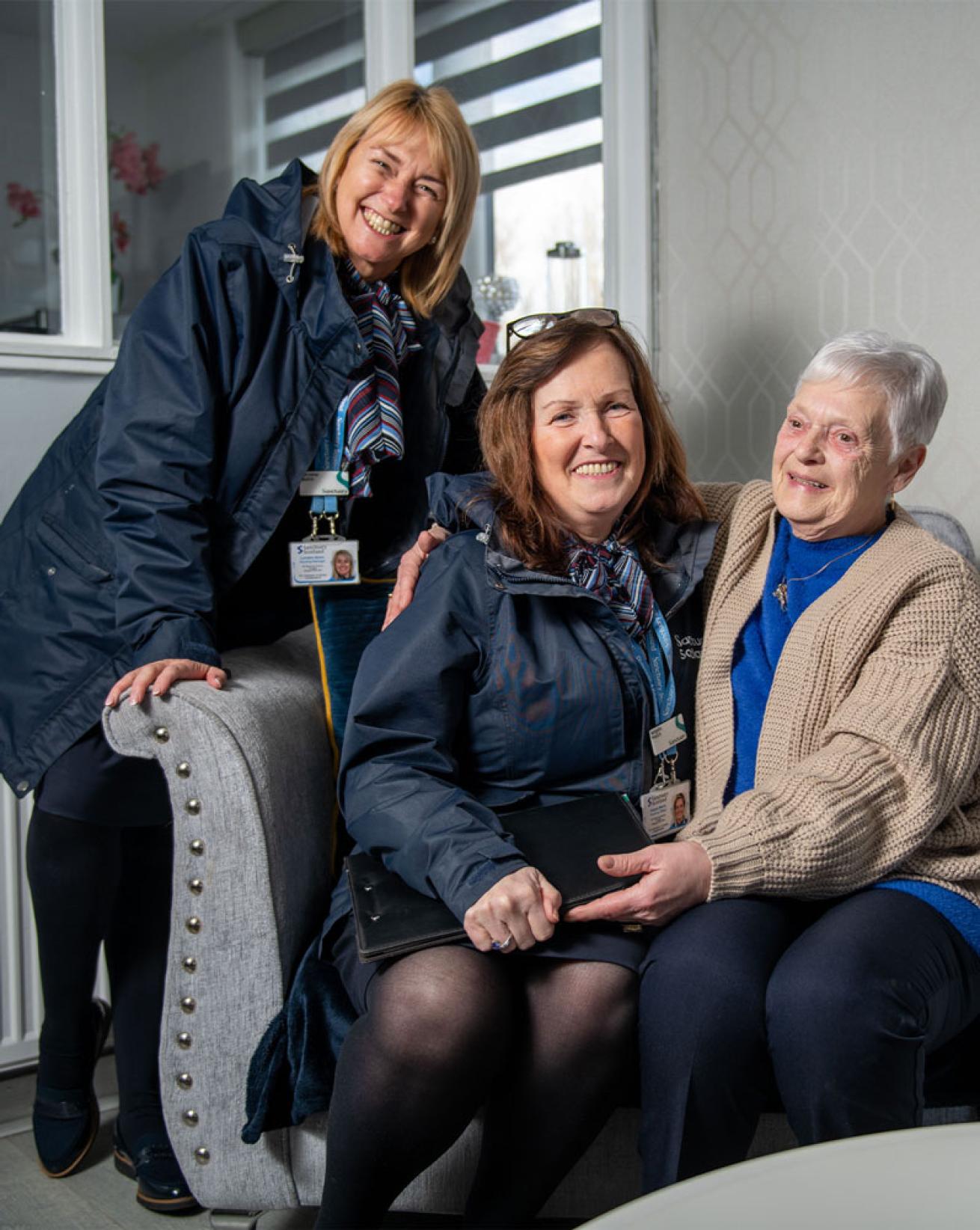 Two Sanctuary Scotland Housing Officers sitting in a living room with an elderly resident who has their arm around one of the housing officers