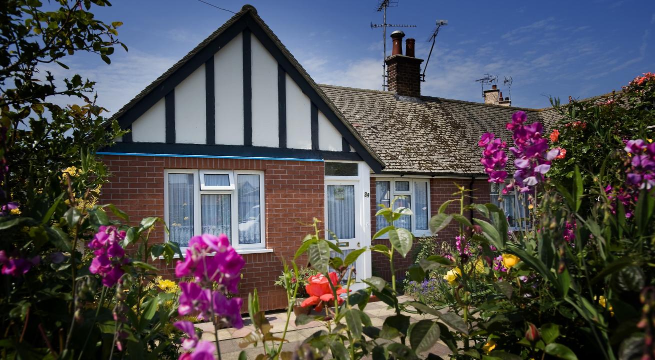 Exterior of a bungalow with red-brick and mock tudor exterior. In the foreground is a collection pink, yellow and red flowers growing out of shrubbery.