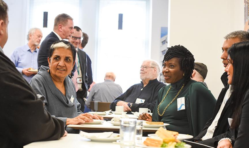 Members of Sanctuary's boards standing and sitting in a canteen with plates of food on the tables.