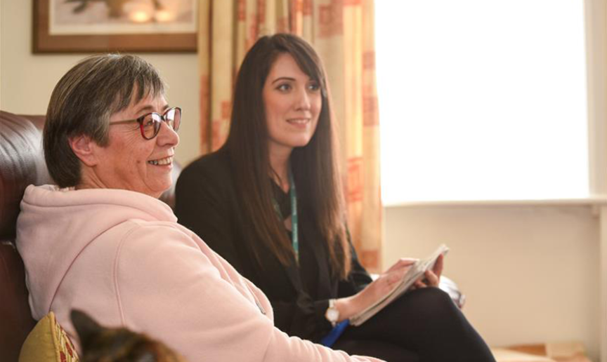 Alt text: A Sanctuary customer, a middle-aged woman with short grey hair and glasses, wearing a light pink hoodie, sits on a brown leather sofa, smiling. Next to her, a Sanctuary Housing Officer, a younger woman with long dark hair, dressed in a black blazer and wearing a lanyard, holds a notepad and pen. They are in a well-lit living room with cream curtains, a framed picture on the wall, and a cat partially visible in the foreground.