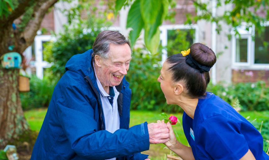 Person in blue jacket holds pink flower while another in blue scrubs leans forward; both are outdoors near a tree and building with greenery.