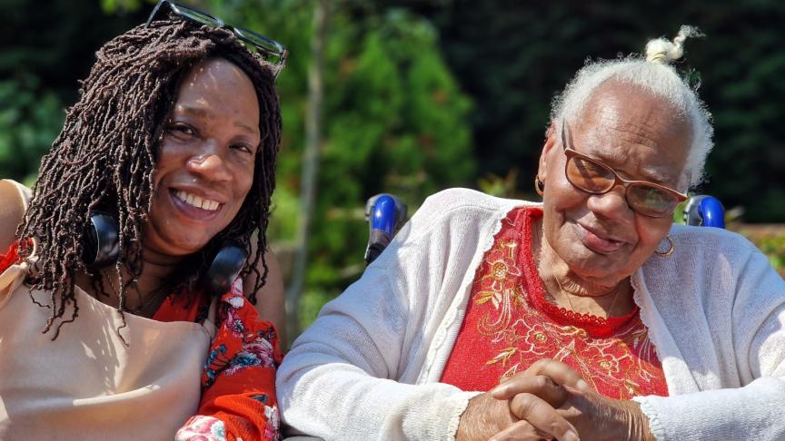 A mother and daughter sat next to each other smiling at the camera, the older woman is sat in a wheelchair