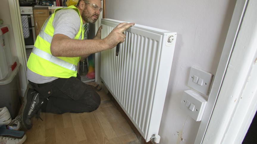 A man wearing a yellow high visibility jacket and protective goggles kneeling in a hallway working on a radiator