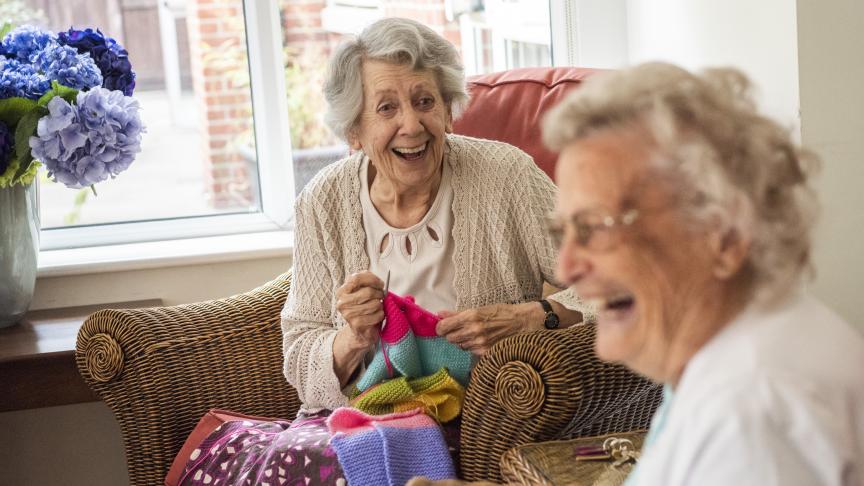 Two elderly women sitting in wicker chairs and laughing whilst knitting with very bright coloured wool