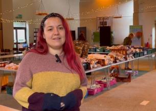 A woman standing in a large room with tables behind her stacked with food and produce