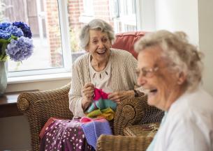 Two elderly women sitting in wicker chairs and laughing whilst knitting with very bright coloured wool