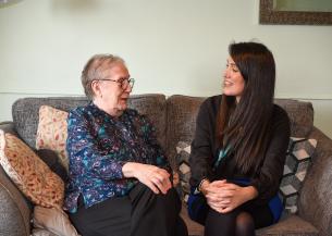 senior woman and a younger woman are sitting together on a cozy gray sofa, smiling and engaging in conversation. The older woman, wearing glasses and a patterned blouse, appears joyful, while the younger woman, dressed in a black top, is laughing. Decorative pillows are arranged on the sofa, and the background features soft lighting and a hint of wall decor, creating a warm and inviting atmosphere.