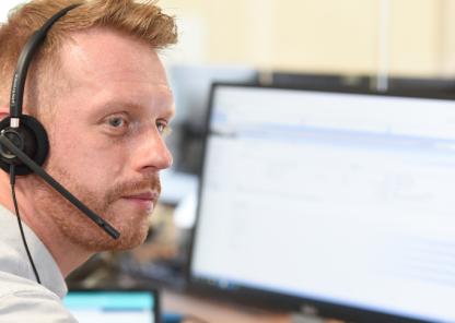 A ginger haired man with a beard wearing a phone headset. There are computer screens on a desk visible in the background.